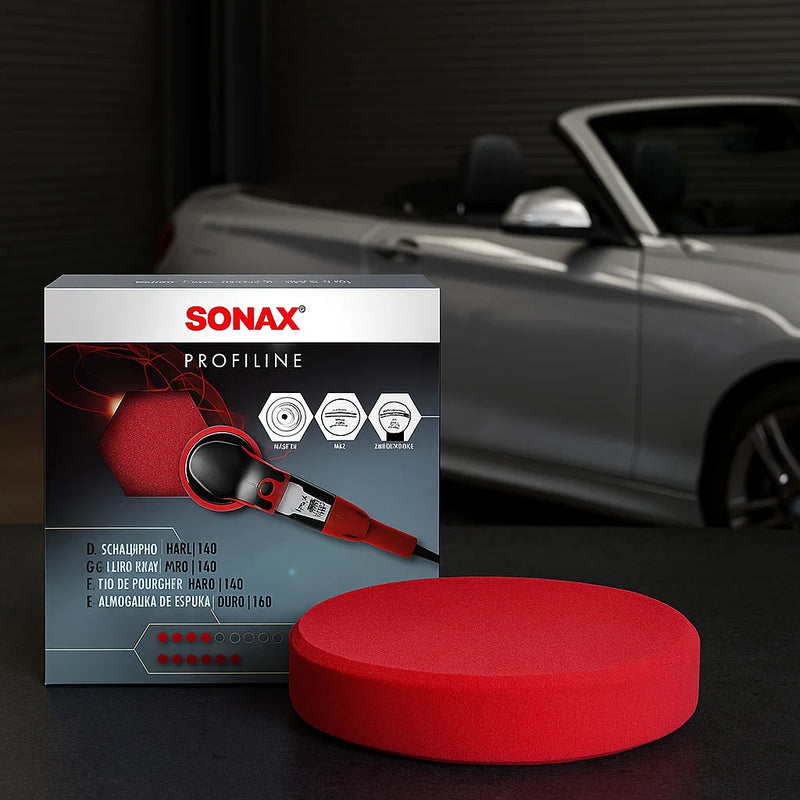 Produktfoto eines roten Sonax Profiline Polierpads (Hard) inklusive Verpackung, aufgenommen in einer Werkstatt mit silbernem Cabrio im Hintergrund