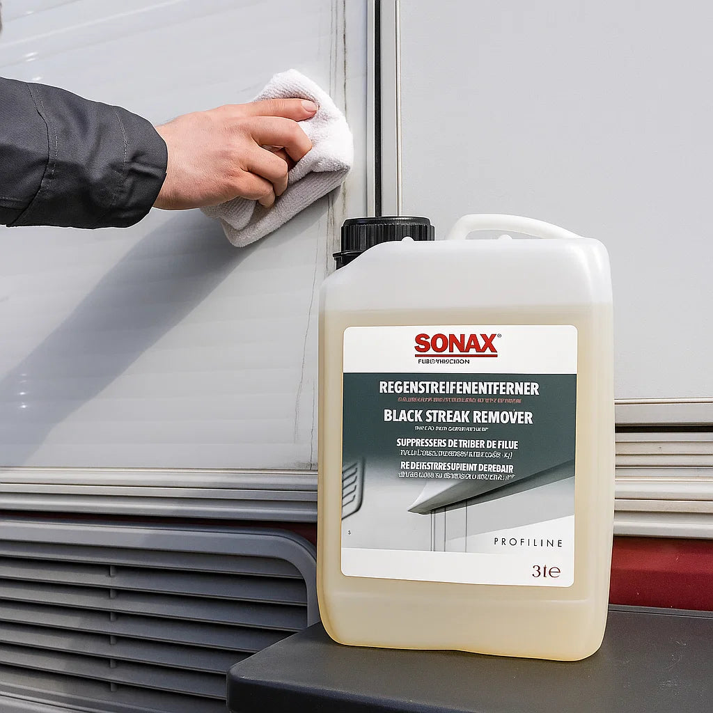 Kanister Sonax Profiline Regenstreifenentferner vor einem Wohnmobil, während eine Hand mit einem Tuch schwarze Regenstreifen von der Außenwand entfernt