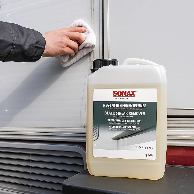 Kanister Sonax Profiline Regenstreifenentferner vor einem Wohnmobil, während eine Hand mit einem Tuch schwarze Regenstreifen von der Außenwand entfernt