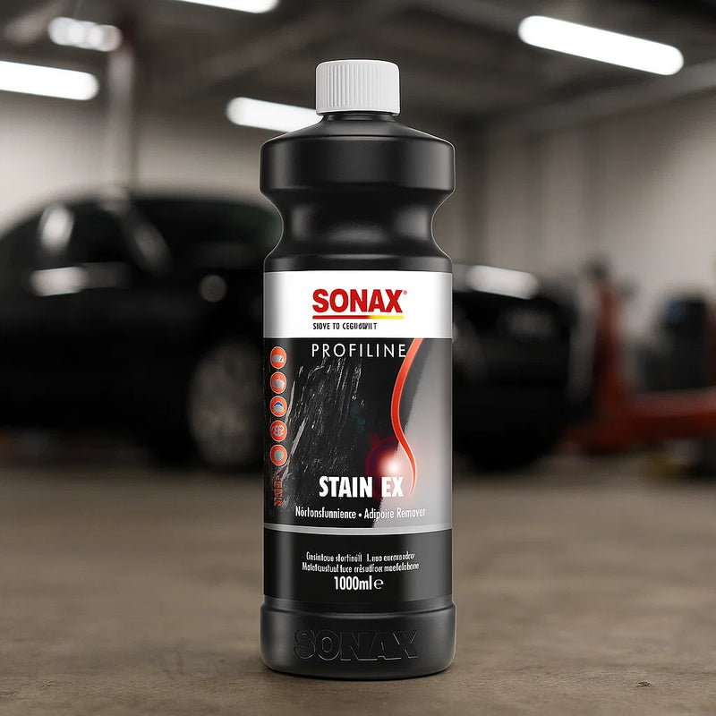 Flasche Sonax Profiline Stain Ex auf dem Werkstattboden mit verschwommenem Fahrzeug im Hintergrund, präsentiert als professioneller Fleckenentferner für Fahrzeuglacke